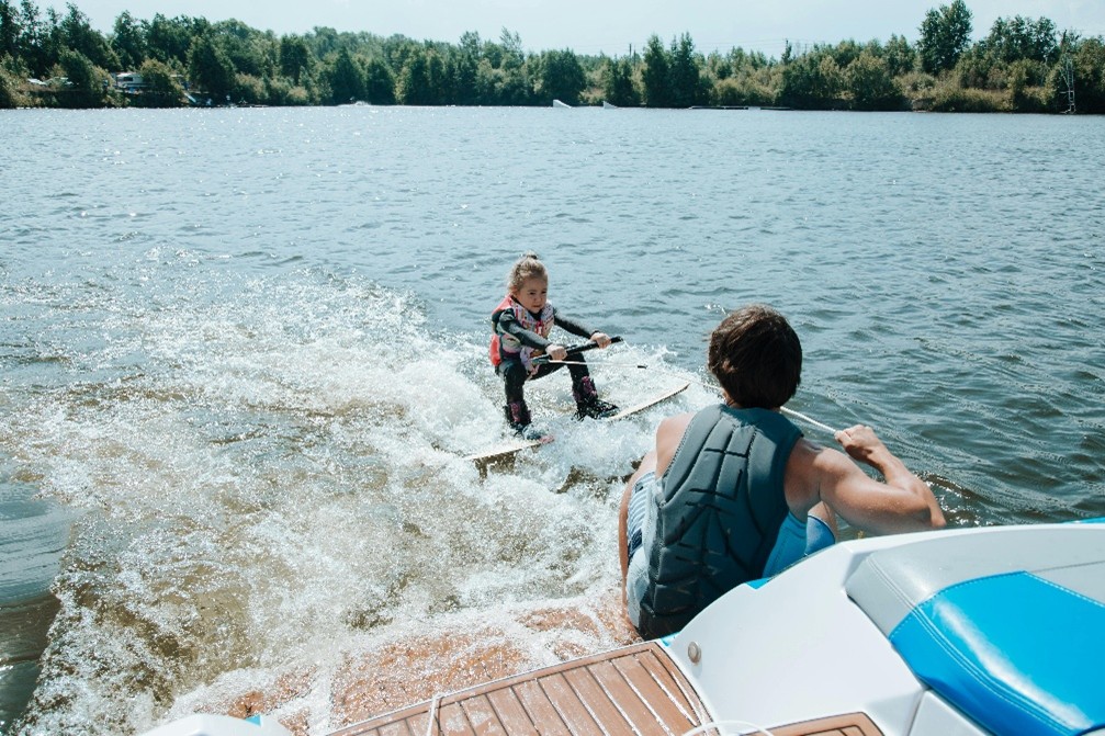 father and daughter wakeboarding on a lake. daughter in the water while father is perched at boat's edge off deck. 