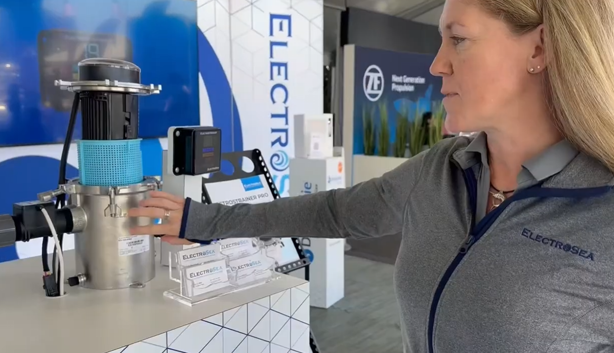 Woman in long-sleeve grey shirt with ElectroSea logo stands in front of a booth display at the Ft Lauderdale boat show and explains technology used in ElectroStrainer. Gestures toward cell canister and the blue sea strainer basket. 